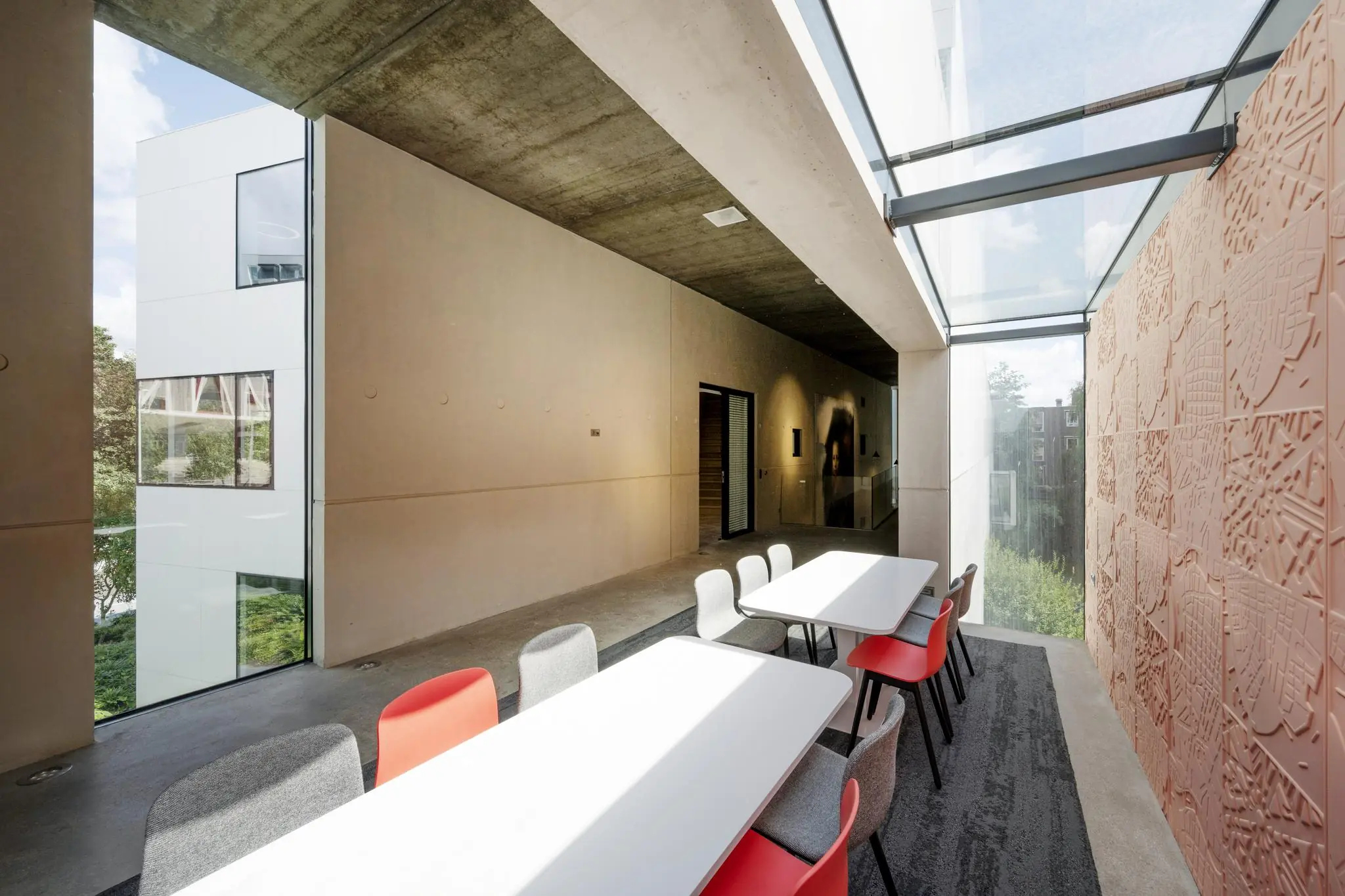 Moderne binnenruimte in de Fred. Roeskestraat met witte tafels, kleurrijke stoelen en een groot raam met uitzicht op een gevel met reliëfpatroon.